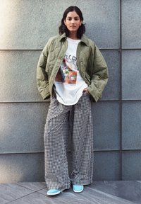 Veste matelassée verte olive sur un t-shirt graphique blanc. Un pantalon à jambes larges en vichy noir et blanc et des baskets bleues complètent la tenue.
