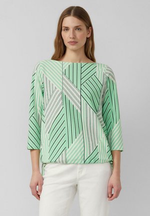 Frau trägt ein mintgrünes und weißes, geometrisch gestreiftes Langarmoberteil kombiniert mit weißen Hosen, steht vor einem schlichten grauen Hintergrund.