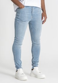 Muž v světlých modrých slim-fit džínách a bílých teniskách, s jednou rukou v kapse, stojící na jednobarevném pozadí.