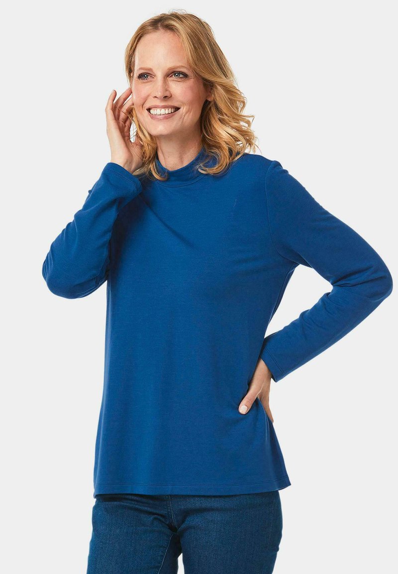 Blaues Langarmoberteil mit hohem Kragen und weicher, glatter Textur. Verfügt über eine lockere Passform und herabgesetzte Schultern, kombiniert mit Jeans.