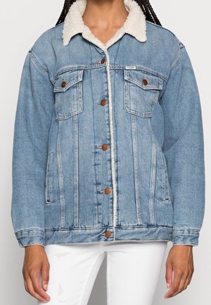 Žena v světle modré džínové bundě s bílým límcem z beránka, hnědými knoflíky a předními kapsami na hrudi, v bílých kalhotách.