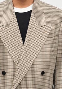 Μπεζ και μαύρο καρό σακάκι με φαρδιές πέτες και μαύρα κουμπιά, φορεμένο πάνω από μαύρο-άσπρο μπλουζάκι με στρογγυλή λαιμόκοψη.