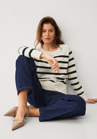 Gebreide trui in crème met zwarte horizontale strepen, ronde halslijn en geribde manchetten, gecombineerd met donkerblauwe wijde jeans en beige puntige hakken.