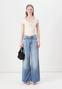 Top entallado de color claro con detalle de nudo, combinado con unos jeans de pierna ancha en azul deslavado. La modelo sostiene un bolso negro pequeño y lleva sandalias beige.