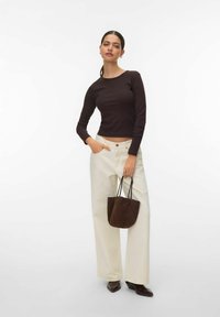 Femme portant un haut à manches longues marron foncé, un pantalon large crème et des bottes foncées, tenant un petit sac à main marron foncé avec des lanières fines.