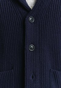 Donkerblauwe ribgebreide cardigan met twee zwarte knopen vastgemaakt over een witte onderhempje, waarbij de bovenste pocketranden zichtbaar zijn.