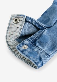 Poignet en denim avec un délavage bleu clair, comportant un intérieur à motifs blancs avec des points rouges et bleus marine, et des fermoirs à bouton-pression en argent.