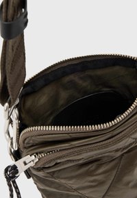 Kleciona torba w kolorze oliwkowym z zamkiem błyskawicznym, z gładką podszewką i czarnym paskiem. Srebrne elementy metalowe podkreślają jej design.