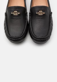 Mocassins Coach en cuir noir avec logo cheval et carrosse doré et texte « Coach New York » sur le dessus, vus de dessus.