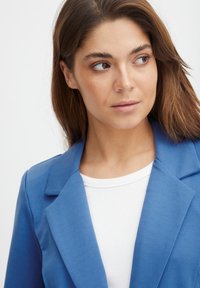Světlomodrý blazer se strukturovaným designem a klopami, v kombinaci s bílým ribovaným topem. Hladká textura, přiléhavý střih, minimální akcenty.
