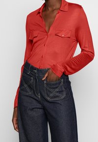 Camicia rossa a maniche lunghe con colletto, due tasche sul petto e chiusura con bottoni; abbinata a pantaloni a vita alta in denim scuro con cuciture a contrasto.