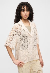 Γυναίκα με σγουρά μαλλιά που φοράει μπεζ πλεκτό τοπ με κοντά μανίκια πάνω από ένα καφέ εσώρουχο και ταιριαστά πλεκτά σορτς, ποζάροντας με το χέρι στη μέση.