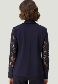 Chaqueta de marina con mangas largas de encaje, silueta ajustada y cuello con muesca. Cuerpo de tela suave, con intrincados detalles de encaje floral en las mangas.