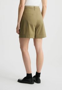 Pantaloni corti khaki realizzati in tessuto leggero, con orlo risvoltato, due tasche posteriori e una cintura texturizzata. Abbinati a scarpe e calze nere.