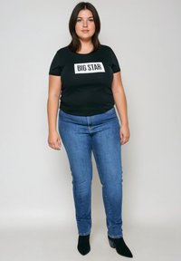 Kobieta stojąca, ubrana w czarną koszulkę z napisem "BIG STAR", niebieskie dżinsy i czarne botki, na jasnym, jednolitym tle.