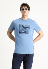 Camiseta de color azul claro con cuello redondo, que presenta un gráfico cuadrado en azul marino con patrones de olas lineales. Mangas cortas, textura suave.