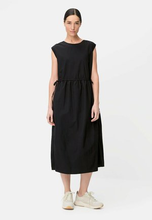 Kvinde står med front mod kamera iført sort ærmeløs midi-kjole med sidesløjfer og hvide sneakers mod en ensfarvet hvid baggrund.