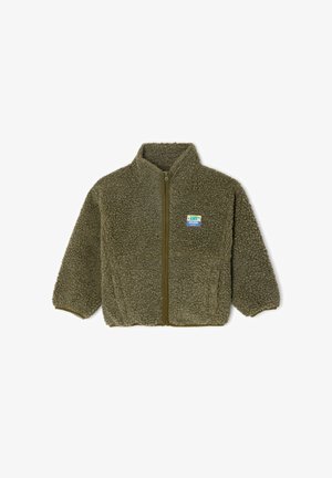 Groen pluizig jack, groene trui, grijze joggingbroek en witte sneakers met bruine accenten. Staat met handen bij de mond, glimlachend.