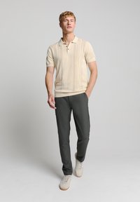 Beige poloshirt met korte mouwen en een gestructureerde voorzijde, gecombineerd met donkergroene slim-fit broeken en lichte sneakers. Eenvoudig, schoon ontwerp.