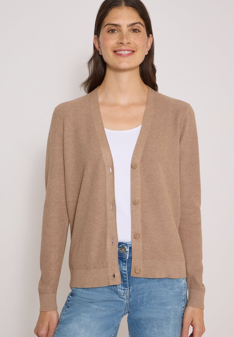 Femme portant un cardigan boutonné marron clair sur un haut blanc et un jean bleu, souriante sur un fond uni.