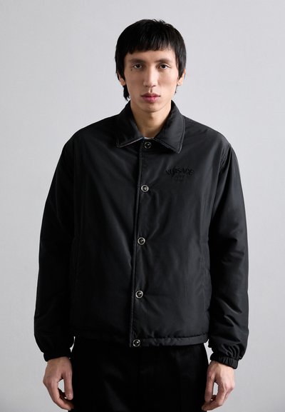 Black padded jacket with a button-up front, stand collar, and ribbed cuffs. Features embroidered logo on the chest and glossy buttons.