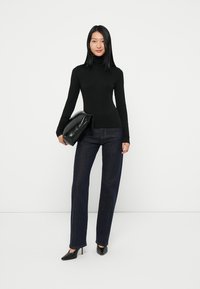 Zwarte coltrui, strak ontwerp, gecombineerd met donkerblauwe rechte jeans. Het model houdt een zwarte leren clutch vast. Zwarte hakken maken de look compleet.