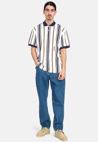 Polo a maniche corte a righe in bianco, blu navy e senape. Presenta un colletto con bottoni e un piccolo logo sul davanti. Indossato con jeans blu.
