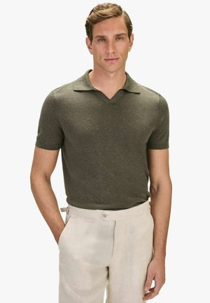 Man in een olijfgroene, korte mouwen, gebreide polo, ingestopt in beige hoge taillebroeken met verstelbare riempjes aan de zijkanten, staand met één hand in de zak.