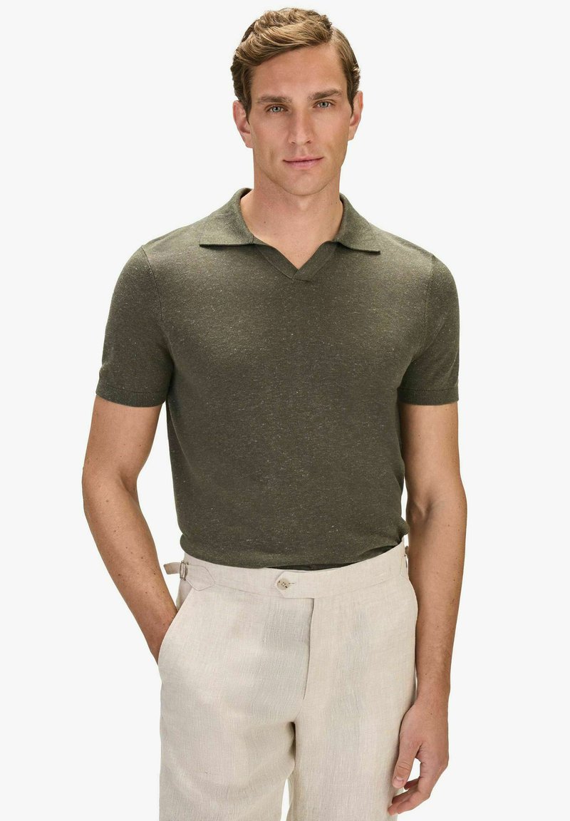 Homme portant un polo en tricot à manches courtes couleur olive rentré dans un pantalon beige taille haute avec des bretelles réglables sur les côtés, debout avec une main dans la poche.