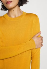 Maglione giallo a maglia con scollatura rotonda e maniche lunghe. Texture liscia e design aderente. La mano del modello poggia sul braccio.