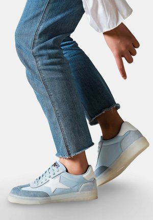 Personne portant des baskets bleu clair avec un détail d'étoile blanche et un jean bleu à ourlet effiloché, pointant vers la chaussure gauche.