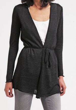 Femme portant un cardigan cache-cœur noir à manches longues noué à la taille par-dessus un top blanc et un pantalon gris clair.