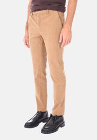Uomo che indossa pantaloni di velluto a coste beige con tasche laterali e scarpe stringate in pelle nera, in piedi di fronte a uno sfondo bianco.