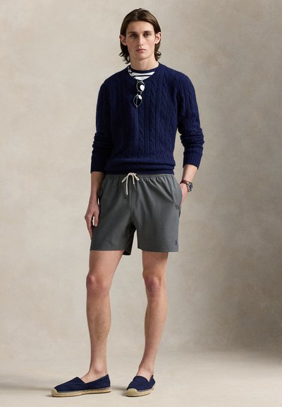 Tamsiai mėlynas megztinis su pynimu, pilkos maudymosi šortai su raišteliu ir tamsiai mėlynos espadrilės. Modelis stovi neutralioje fone.