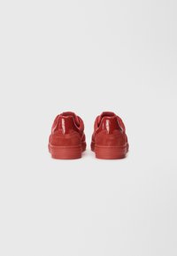 Sneaker rosse in suede con tacco rosso lucido, upper testurizzato, colletto imbottito e suola in gomma. La vista posteriore evidenzia i dettagli delle cuciture.