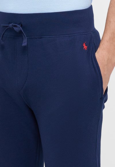 Jogging en maille bleu marine avec taille élastique, dotés de poches latérales et d'un petit logo rouge sur la hanche gauche. Tissu texturé.