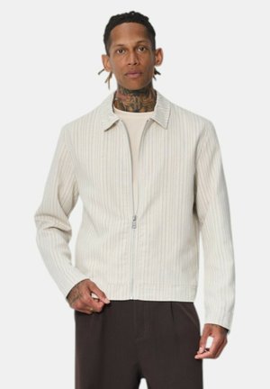 Homme avec des tatouages portant une veste zippée beige clair à fines rayures et un pantalon foncé, debout devant un fond uni.