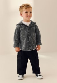 Giacca di jeans grigia con cappuccio e motivo sottile, abbinata a pantaloni cargo neri e sneakers nere. Outfit casual e a strati per bambini.