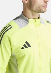 Neon-gelbe Sportjacke mit Reißverschluss, grauen Akzenten am Kragen und schwarzen vertikalen Streifen an den Ärmeln, versehen mit einem Logo.