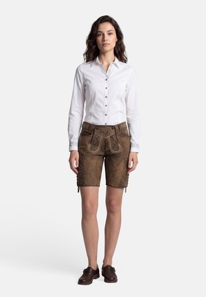 Frau steht frontal, trägt ein weißes Hemd mit Knöpfen, braune, bestickte Lederhosen und dunkle Loafers vor hellem Hintergrund.