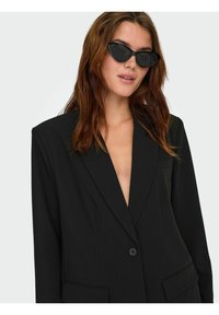 Sort blazer med dyb V-udskæring, strukturerede skuldre, to forlommer og en enkelt knaplukning. Model bærer cat-eye solbriller.