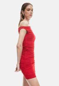Robe rouge sans manches avec une silhouette ajustée, des détails froncés et une longueur au genou. Tissu lisse et extensible.