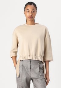 Felpa beige corta con scollatura tonda, maniche a tre quarti e vita elasticizzata. Abbinata a pantaloni a quadri grigi.