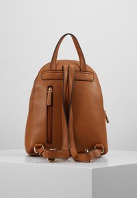 Sac à dos en cuir marron avec deux bretelles réglables, une poignée supérieure et une poche vertical zippée sur un piédestal blanc.