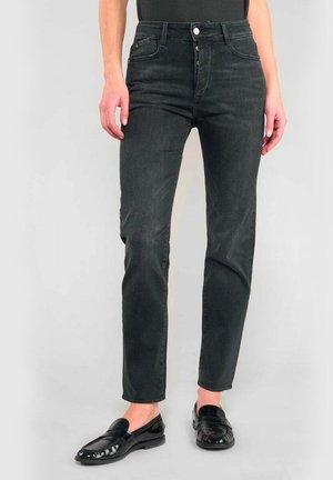Donna che indossa jeans neri a vita alta con chiusura a bottoni e mocassini neri lucidi, in piedi di fronte a uno sfondo chiaro e semplice.