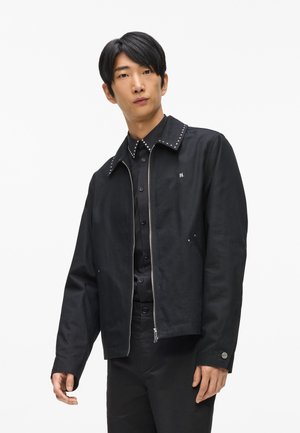 Black jacket with a studded collar, zip front, and side pockets. Made from textured fabric, featuring a simple logo on the chest.