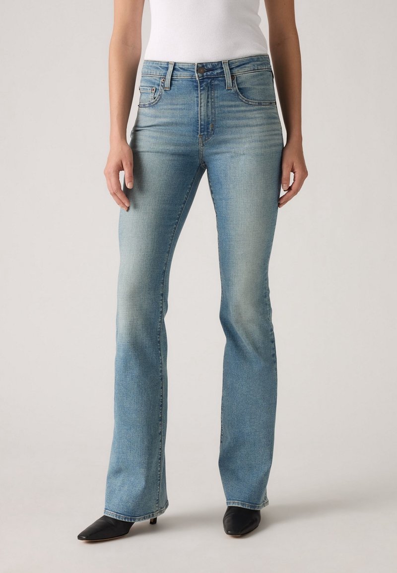 Femme portant un jean bootcut taille haute bleu clair et des chaussures à talons noires à bout pointu, debout avec les bras détendus le long du corps.
