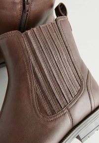 Botas de tobillo de cuero marrón con un diseño acanalado en los lados, cierre de cremallera y un acabado texturizado. Suela resistente con detalles de tracción.