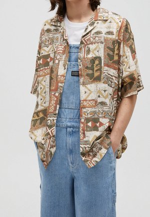 Personne portant une chemise à manches courtes à motifs par-dessus un t-shirt blanc et une salopette en denim bleu, les mains dans les poches.