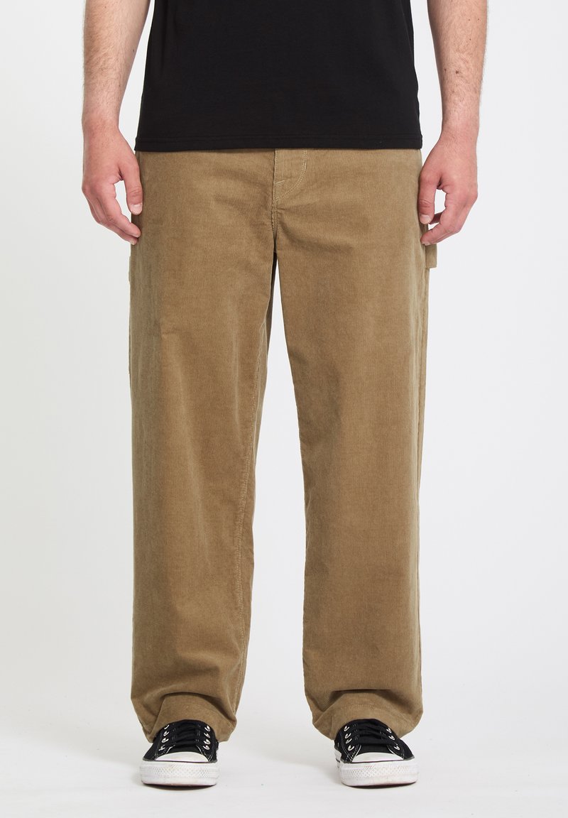 Mies, joka pukeutunut ruskeisiin pusakkaiset cordurohousuihin, mustiin lenkkareihin valkoisilla pohjilla ja mustaan t-paitaan, seisoo yksinkertaisen valkoisen taustan edessä.